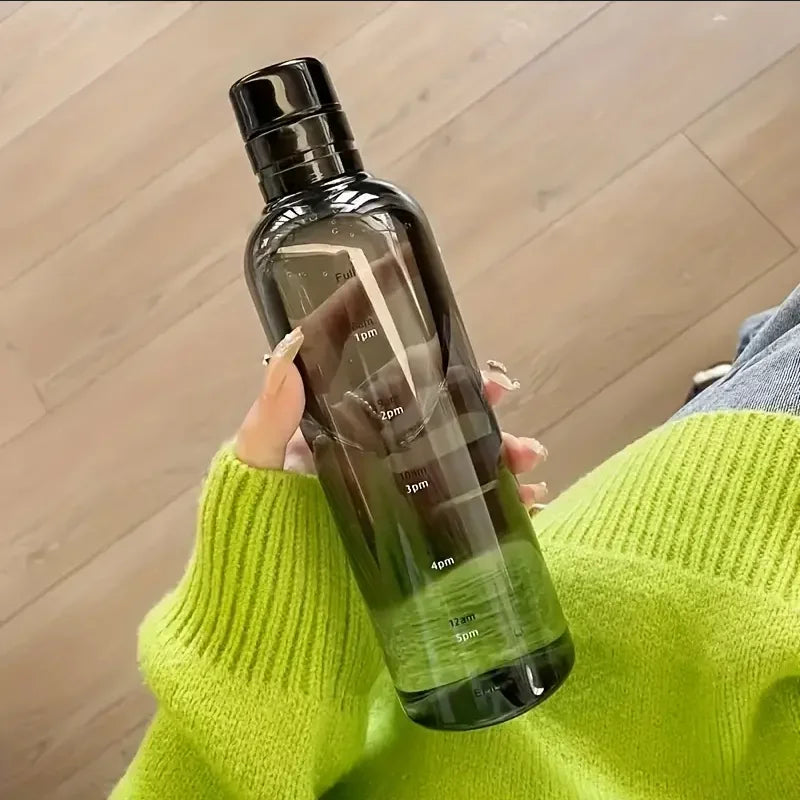 Hand holding a black water bottle with time markers, person in green sweater, wood floor